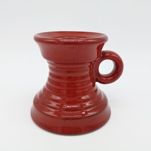 May include: A red ceramic candle holder with a handle. The holder has a wide base and a narrow top.