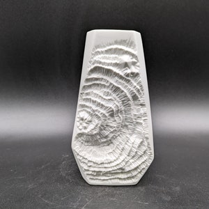 May include: White ceramic mould with a textured design resembling a tree trunk. The mould is shaped like a vase and has a smooth, rounded base.