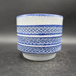 May include: A ceramic planter with a cylindrical shape. The planter features a textured, light blue and white upper section, with two bands of intricate blue woven-like patterns. The base is a solid, light blue color.