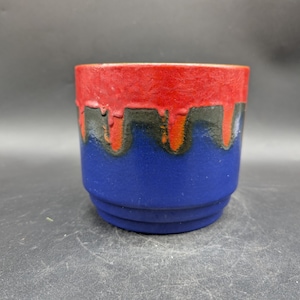 Könnte beinhalten: Eine Keramiktasse mit einem geschichteten Design. Der obere Teil ist rot, gefolgt von einem schwarzen und orangefarbenen Band, und die Basis ist blau. Die Tasse hat eine strukturierte Oberfläche und eine zylindrische Form.