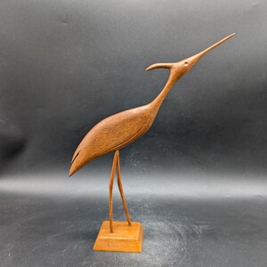 May include: A wooden sculpture of a bird with a long neck and beak, standing on two thin legs attached to a square base. The bird is a warm brown color, and the sculpture has a smooth finish.