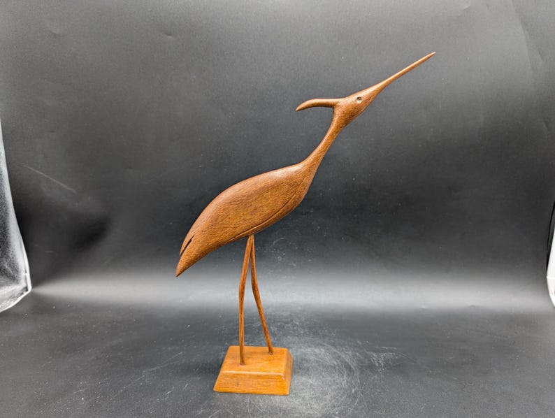 May include: A wooden sculpture of a bird with a long neck and beak, standing on two thin legs attached to a square base. The bird is a warm brown color, and the sculpture has a smooth finish.