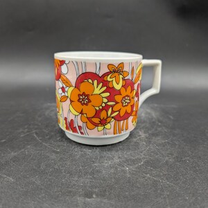 Könnte beinhalten: Weiße Keramiktasse mit Retro-Blumenmuster in Orange, Gelb und Rot auf rosa Hintergrund. Die Tasse hat einen quadratischen Henkel und einen leicht ausgestellten Boden, ideal für Kaffee oder Tee.