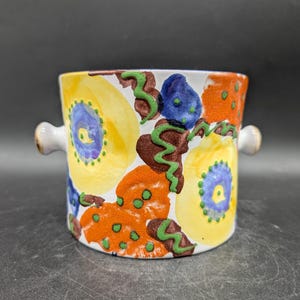May include: A ceramic container with a vibrant floral design. The hand-painted pattern features large yellow, orange, and blue flower-like shapes with green accents. It has two small, white handles and is suitable for various uses.
