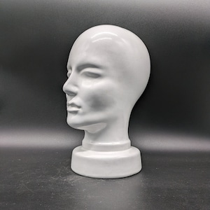 Könnte beinhalten: Ein weißer Keramik-Mannequin-Kopf mit glatter, matter Oberfläche. Der Kopf ist leicht zur Seite geneigt und die Augen sind geschlossen. Der Mannequin steht auf einem weißen Sockel.