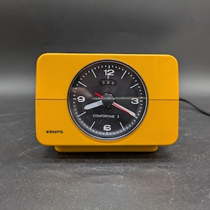 Könnte beinhalten: Ein gelber Wecker mit schwarzem Zifferblatt und weißen Zahlen. Die Uhr zeigt "Krup's" und "Comfortime 3".
