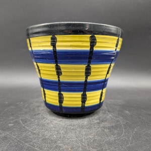 Könnte beinhalten: Eine Keramikschale mit schwarzem Rand und Boden, mit horizontalen gelben und blauen Bändern. Vertikale schwarze Linien mit kleinen rechteckigen Formen verlaufen die Seiten hinunter. Die Schale hat eine leicht ausgestellte Form.