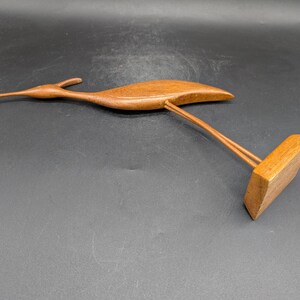 May include: A wooden bird sculpture with a long beak and outstretched wings. The bird is perched on a rectangular base, supported by two thin wooden rods. The sculpture is made of a warm, medium-toned wood.