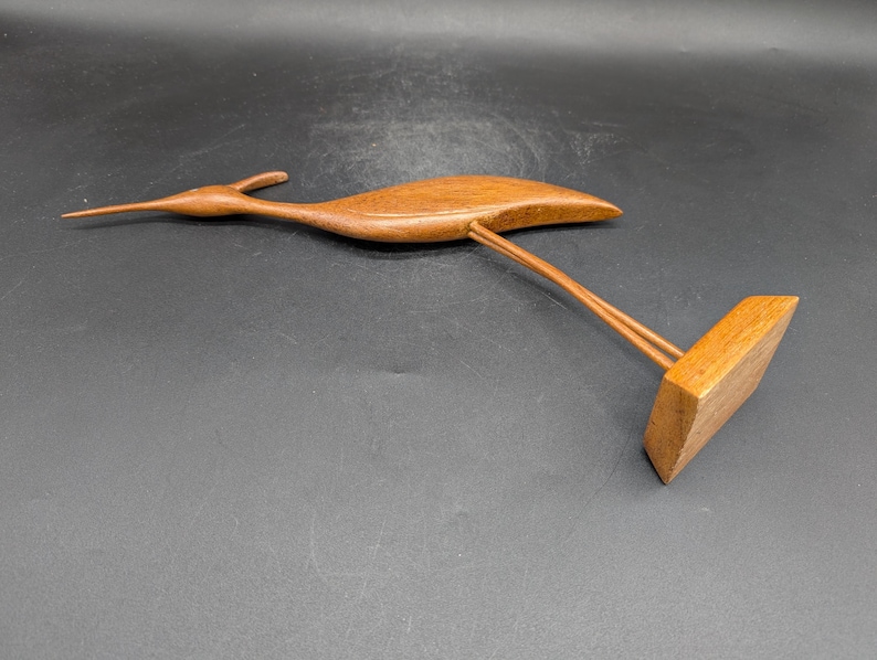 May include: A wooden bird sculpture with a long beak and outstretched wings. The bird is perched on a rectangular base, supported by two thin wooden rods. The sculpture is made of a warm, medium-toned wood.