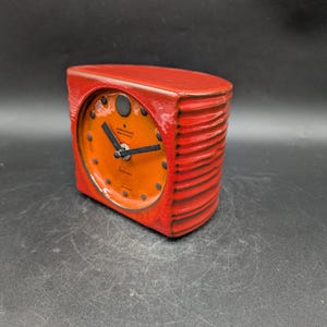 Könnte beinhalten: Ein roter Vintage-Wecker mit orangefarbenem Zifferblatt und schwarzen Zeigern. Die Uhr hat eine rechteckige Form mit geriffelter Textur an den Seiten. Das Zifferblatt hat schwarze Punkte als Stundenmarkierungen.