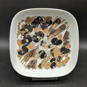 Può includere: Un piatto quadrato in ceramica bianca con un motivo floreale decorativo. Il design presenta forme astratte nere, marroni e dorate con accenti arancioni. Il piatto ha angoli arrotondati e un bordo leggermente rialzato.