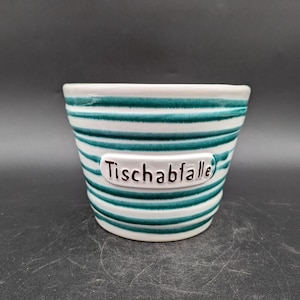 Puede incluir: Pequeño cuenco de cerámica blanca con rayas horizontales turquesas. Tiene una etiqueta blanca con la palabra "Tischabfalle" en letras negras. Probablemente se utiliza con fines decorativos o como maceta pequeña.