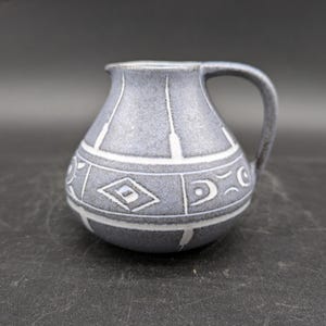 May include: A small, gray ceramic pitcher with a white handle and geometric designs. The pitcher has a rounded body and a slightly flared lip.