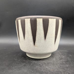 Op de afbeelding: Een keramische kom met een lichtgrijze buitenkant en een donkerbruine binnenkant. De kom heeft een herhalend patroon van donkerbruine, naar beneden wijzende driehoeken tegen een lichtgrijze achtergrond. De kom heeft een afgeronde vorm en een kleine, cirkelvormige basis.