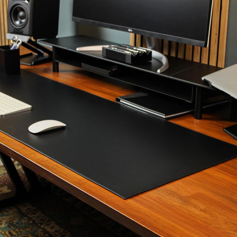 May include: A black leather desk mat on a wooden desk. The mat is large enough to cover most of the desk surface. A white computer mouse is on the mat.