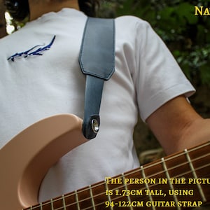May include: A person wearing a white t-shirt with a blue shark graphic is holding a light pink electric guitar with a black guitar strap. The strap is adjustable from 94-122cm. The text "NAVY" is in the upper right corner of the image. The text "THE PERSON IN THE PICTURE IS 1.73CM TALL, USING 94-122CM GUITAR STRAP" is at the bottom of the image.
