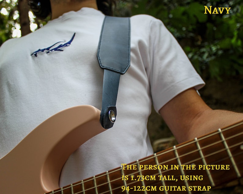 May include: A person wearing a white t-shirt with a blue shark graphic is holding a light pink electric guitar with a black guitar strap. The strap is adjustable from 94-122cm. The text "NAVY" is in the upper right corner of the image. The text "THE PERSON IN THE PICTURE IS 1.73CM TALL, USING 94-122CM GUITAR STRAP" is at the bottom of the image.