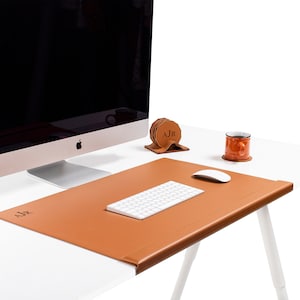 Könnte beinhalten: Eine braune Leder-Schreibtischunterlage mit einem personalisierten Monogramm "AJR" und einem genähten Rand. Die Unterlage liegt auf einem weißen Schreibtisch mit einem Computermonitor, einer Tastatur, einer Maus und einer Tasse.