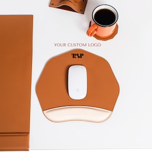 Può includere: Un tappetino per mouse in pelle marrone con un poggiapolsi bianco e un mouse wireless bianco. Il tappetino per mouse ha una forma esagonale e il testo "YOUR CUSTOM LOGO" e "T&P" stampato su di esso.