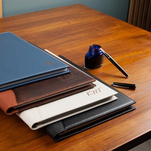 May include: A selection of personalised leather journals in blue, brown, white, and black, each with initials. A blue ink bottle and a black pen sit on a wooden surface, suggesting a stationery theme.