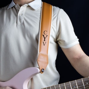 May include: A brown leather guitar strap with the initials "CYV" engraved on it. The strap is worn by a person holding a pink electric guitar.