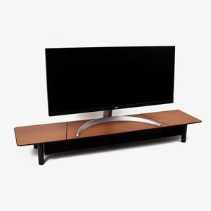 May include: A brown monitor stand with a black base and legs supports a large black computer monitor. The stand has a rectangular shape with a smooth, leather-like surface. The monitor has a silver stand.