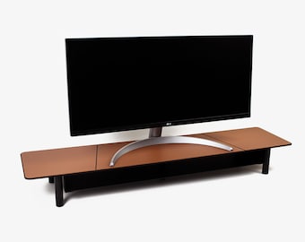 May include: A brown monitor stand with a black base and legs supports a large black computer monitor. The stand has a rectangular shape with a smooth, leather-like surface. The monitor has a silver stand.