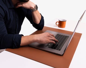May include: A person working on a silver laptop computer with a brown leather desk mat. A cup of coffee sits on the desk to the right of the laptop.