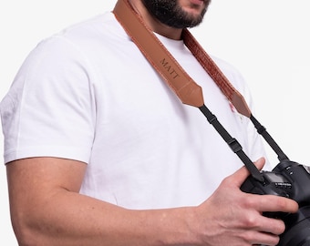 Może przedstawiać: Brązowy skórzany pasek do aparatu fotograficznego z wygrawerowanym imieniem "MATT". Pasek jest noszony na ramieniu osoby trzymającej czarny aparat.