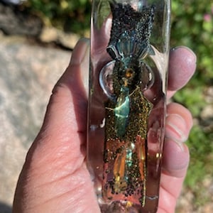 Könnte beinhalten: Ein rechteckiger, klarer Gegenstand aus Harz mit eingebetteten irisierenden und glitzernden Elementen. Das Objekt hat ein abgerundetes, blasenartiges Merkmal in der Mitte. Das Harz umschließt eine Mischung aus Farben, darunter Gold, Grün und Schwarz, wodurch ein einzigartiges, dekoratives Stück entsteht.