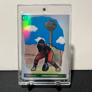 May include: A Downtown Raiders softball trading card in a clear protective case. The card features an illustration of a softball player in a red and black uniform, with a water tower and clouds in the background. The card is displayed on a black stand.