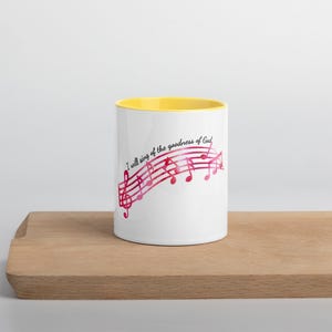 Könnte beinhalten: Weiße Keramiktasse mit gelbem Innenraum, mit einem Notenlinien-Design in Rosa- und Rottönen. Der Text "I will sing of the goodness of God" ist auf die Tasse gedruckt. Die Tasse steht auf einer Holzoberfläche.