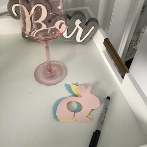 May include: A pink wine glass with the word "Bar" printed on it, a wooden sign that says "Bar", a pink bunny-shaped coaster, and a black Sharpie marker are on a white tray.
