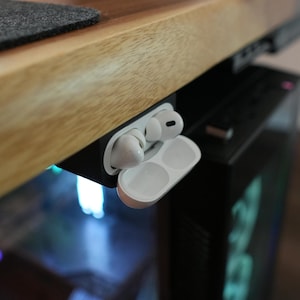 May include: White wireless earbuds and charging case, partially open, mounted under a wooden desk. The earbuds are in the case, and the case is open, revealing the charging slots. The desk has a light wood grain finish.
