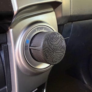 May include: A close-up of a car's temperature control knob with a black, textured knob cover. The knob is labeled "TEMP" and has a blue "MAX A/C" indicator.