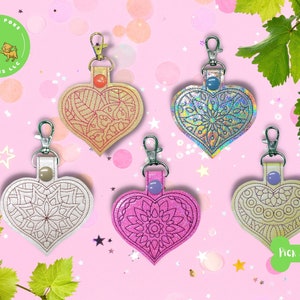 May include: Five heart-shaped keychains with different colors and designs. The keychains are made of leather and have a mandala pattern. The keychains are hanging from a pink background with green leaves.