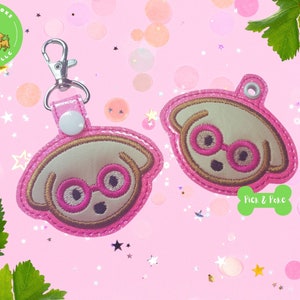 May include: Two pink and gold dog keychains with glasses. The keychains are on a pink background with glitter. The text "Pick & Poke" is on one of the keychains.