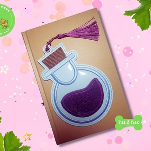 May include: Purple and blue embroidered potion bottle bookmark with a purple tassel. The bookmark is on a brown surface with a pink background.