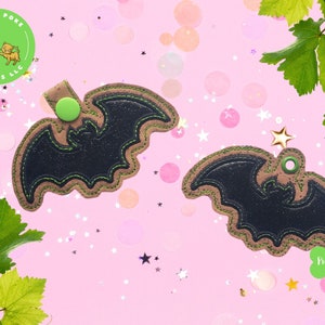 May include: Two black and green bat-shaped keychains with a glittery finish. The keychains are on a pink background with green leaves and silver stars.