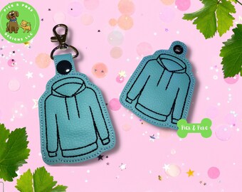 May include: Two teal blue fabric keyrings shaped like hoodies. The keyrings are embroidered with black stitching. The keyrings are on a pink background with silver glitter.