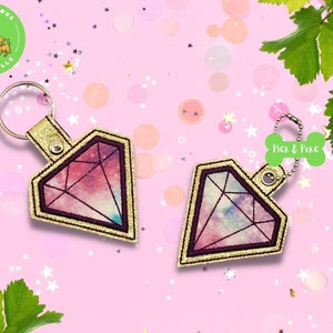 May include: Two diamond-shaped keychains with a galaxy-like design. The keychains are pink, purple, and blue with gold accents. The keychains are on a pink background with white stars.