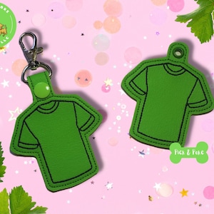 May include: Green felt keychain shaped like a t-shirt with black stitching. The keychain has a silver metal ring and a green button. The keychain is on a pink background with green leaves and silver glitter.