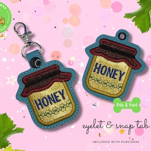 Op de afbeelding: Sleutelhanger en tag set met honingpot. Het honingpotontwerp heeft een goudglitter centrum met het woord "HONEY" in marineblauw. Het deksel is bruin met een decoratief patroon en de rand is teal. Eén sleutelhanger heeft een zilveren sluiting.