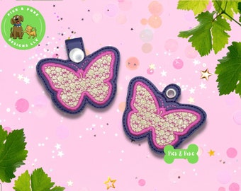 Puede incluir: Dos llaveros en forma de mariposa rosa y blanca con un acabado brillante. Los llaveros están hechos de tela y tienen un borde azul. Los llaveros están sobre un fondo rosa con lunares blancos.