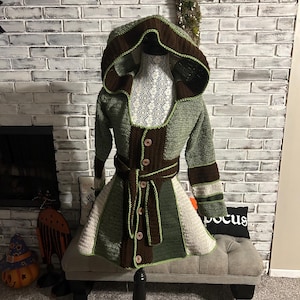 May include: A handmade crochet coat with a hood, displayed on a mannequin. The coat features a color block design in shades of green, brown, and cream, with wooden buttons and a matching belt. The coat is displayed on a black stand.