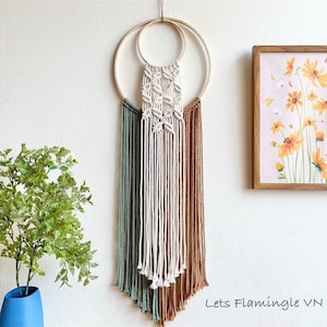 May include: Macrame wall hanging with a geometric design, featuring a cream-coloured central section and ombre shades of green and brown. The artwork is suspended from two wooden rings. A framed floral print and a potted plant are also visible.