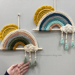 May include: Two macrame rainbow wall hangings featuring mustard yellow fringe, multi-colored yarn arches, and a braided cloud with three dangling raindrops. Each piece is suspended from a horizontal wooden dowel with a hanging cord.