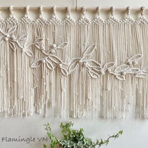 May include: An off-white macrame wall hanging suspended from a wooden dowel, featuring a decorative pattern of knotted leaves and vines across the center, with long, dangling fringe strands ending in small knots. The text Lets Flamingle VN is visible.
