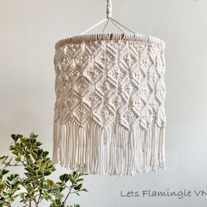 Op de afbeelding: Een witte macramé lampenkap met een geometrisch patroon en franjes. De lampenkap hangt aan een wit plafond.