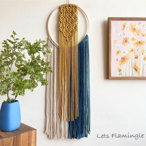 May include: A macrame wall hanging with a circular wooden hoop and white, yellow, and blue yarn. The wall hanging is hanging on a white wall with a framed print of yellow flowers on a pink background.
